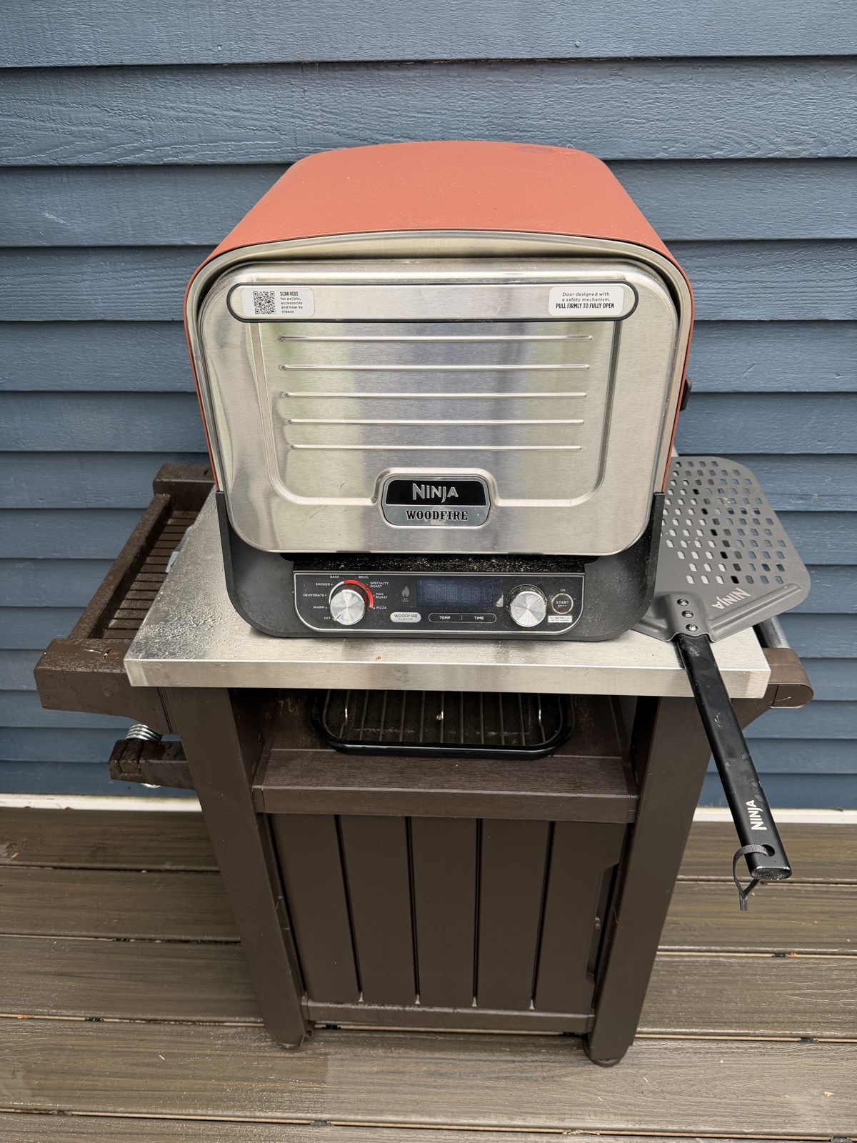 Ninja Woodfire OO101 in terracotta red on a Keter cart against blue siding, with the Ninja perforated peel hanging on the side